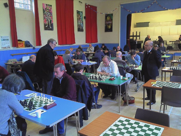 Ein großer Saal mit zwei Personen, die an einem Tisch Schach spielen, mit Münzen auf dem Schachbrett, Vorhängen, Postern, einem Fenster und einem Kalender an der Wand.