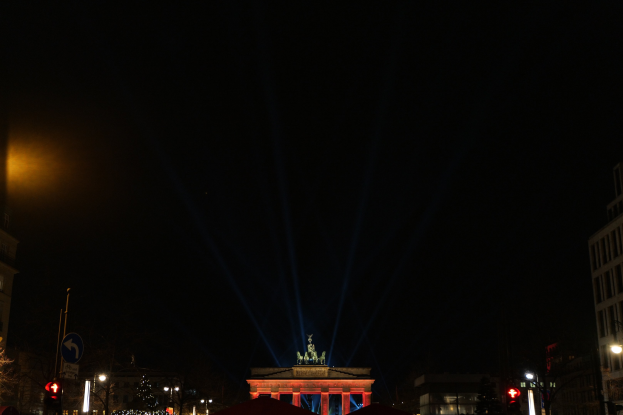 Das Brandenburger Tor in Berlin nachts, von Straßenlaternen beleuchtet, umgeben von Gebäuden, Bäumen, Pfählen, Verkehrsampeln, Schildern und Zelten unter einem dunklen Himmel.