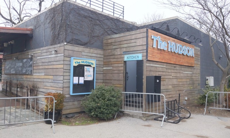 Außenansicht des Hudson-Restaurants in New York City, mit einer Tür, einem Schild, Topfpflanzen, einem Metallzaun, Bäumen, einem bewölkten Himmel und einem Müllbehälter im Vordergrund.