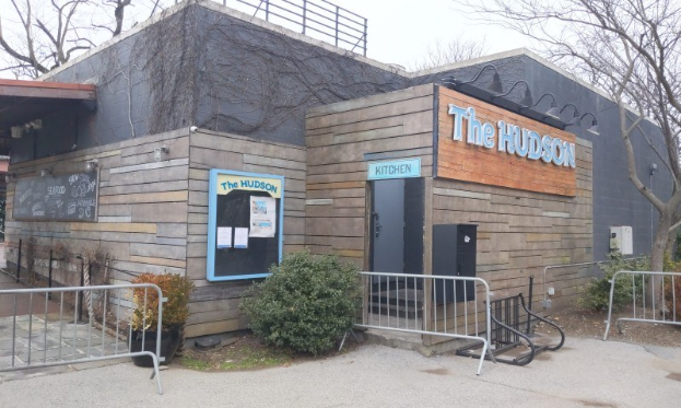 Außenansicht des Hudson-Restaurants in New York City, mit einer Tür, einem Schild, Topfpflanzen, einem Metallzaun, Bäumen, einem bewölkten Himmel und einem Müllbehälter im Vordergrund.