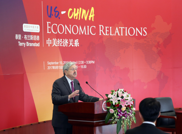 Ein Mann in einem schwarzen Blazer, weißem Hemd und lila Krawatte steht an einem Podium mit zwei Mikrofonen und einem Blumenstrauß, während er vor einer roten Wand spricht, auf der 'US-China Economic Relations' steht, mit einer Person rechts neben ihm sitzend.