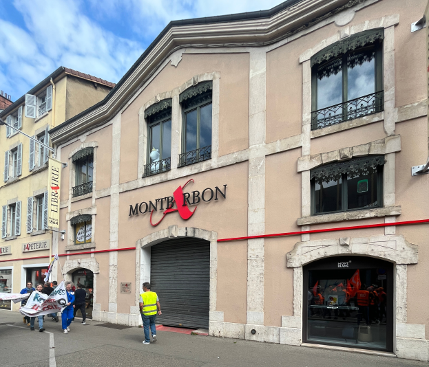 Eine Gruppe von Menschen mit Fahnen geht vor einem Gebäude mit einem Schild vorbei, auf dem 'Montbron' steht, einem Straßenpfahl und einer Fahne mit Bildern und Text unter einem bewölkten Himmel.