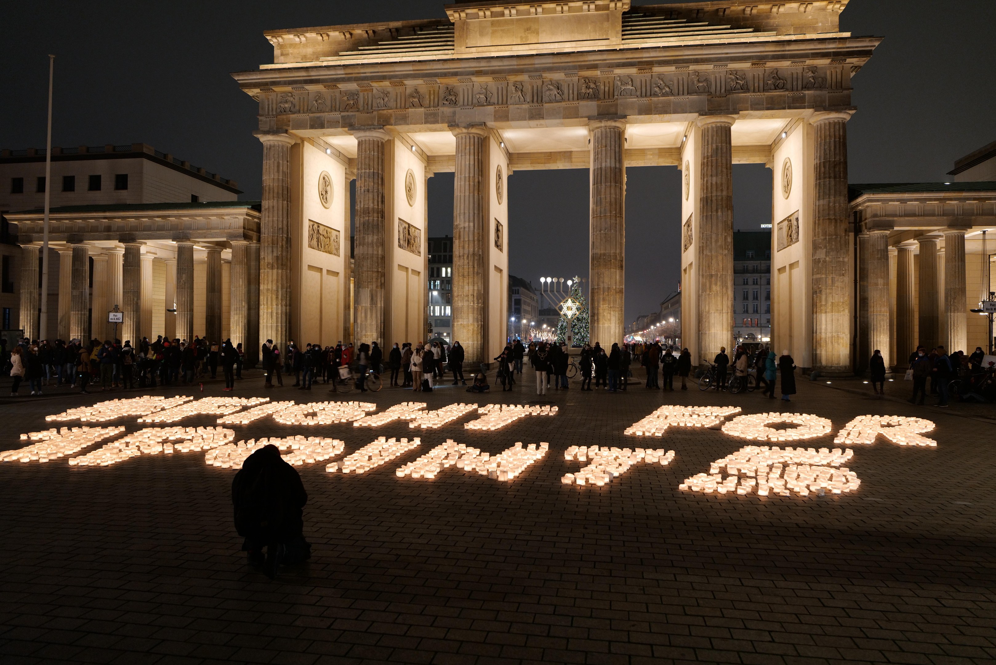 Eine Gruppe von Menschen steht vor dem beleuchteten Brandenburger Tor in Berlin, Deutschland, umgeben von Gebäuden, Pfählen und Lichtern, und die Wörter 'Kampf für die Freiheit' sind auf den Boden geschrieben.