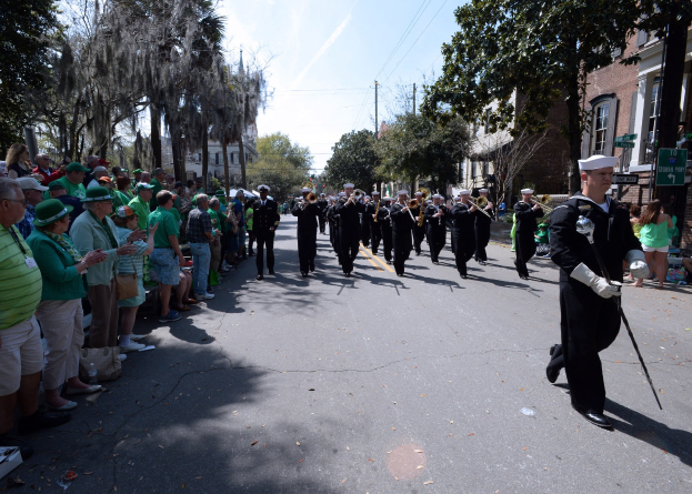 Eine Gruppe von Menschen in Mützen, einige mit Musikinstrumenten, marschiert auf einer Straße während eines St. Patrick's Day-Umzugs, mit Zuschauern auf der linken Seite und einem Gebäude, Bäumen und Strommasten im Hintergrund.