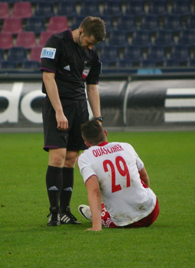 Ein Fußballspieler und Schiedsrichter sitzen auf dem Boden in einem Stadion, beide tragen Sportkleidung.