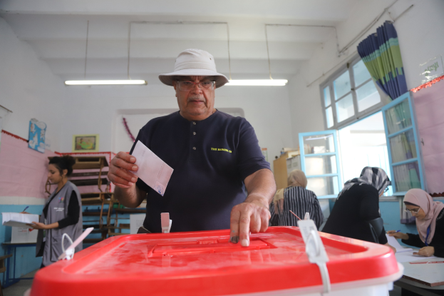 Ein Mann mit Hut stimmt in einer Wahllokal ab, vor einer roten Wahlurne stehend, mit einem Zettel in der Hand, während andere an Tischen sitzen und schreiben, mit einem Fenster und Vorhang auf der rechten Seite.