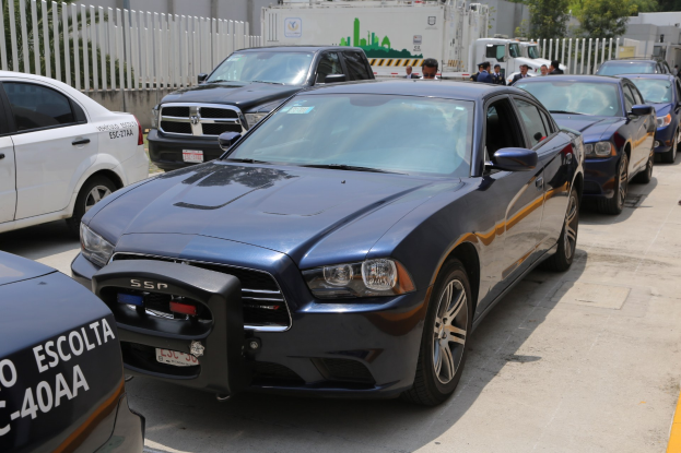 Polizeifahrzeuge auf einem Parkplatz mit Menschen auf der Straße, Zaun, Bäumen, Gebäuden und einem klaren blauen Himmel im Hintergrund.