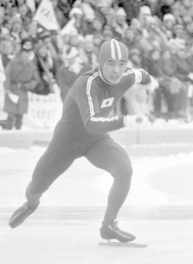 Ein Schwarz-Weiß-Foto eines Eisschnellläufers in Aktion auf dem Eis, der einen Helm und Schlittschuhe trägt, mit ein paar Menschen im Hintergrund, die Fahnen halten.