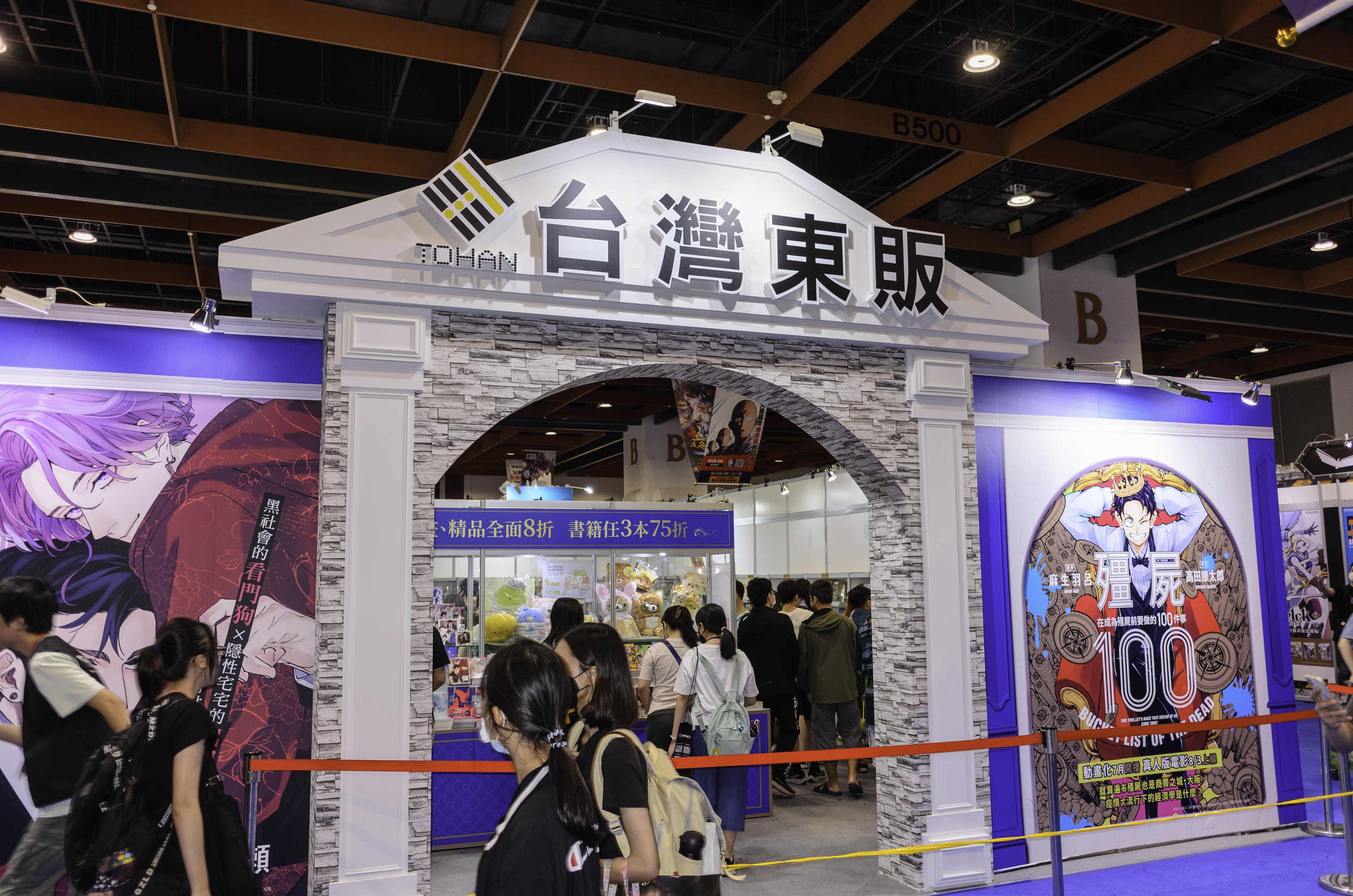 Eine Gruppe von Menschen steht vor einer Spielshow-Bühne auf einer Messe, einige tragen Taschen; die Bühne zeigt Text- und Bildtafeln sowie Deckenlampen.