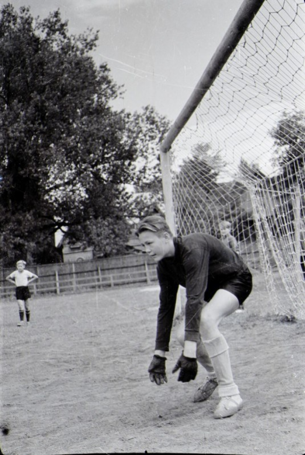 Ein Schwarz-Weiß-Foto eines jungen Jungen, der auf einem Feld Fußball spielt, mit einem Zaun, Bäumen und einem klaren Himmel im Hintergrund, anderen Kindern, die in der Ferne spielen, und einem Schuhwerk im Vordergrund.