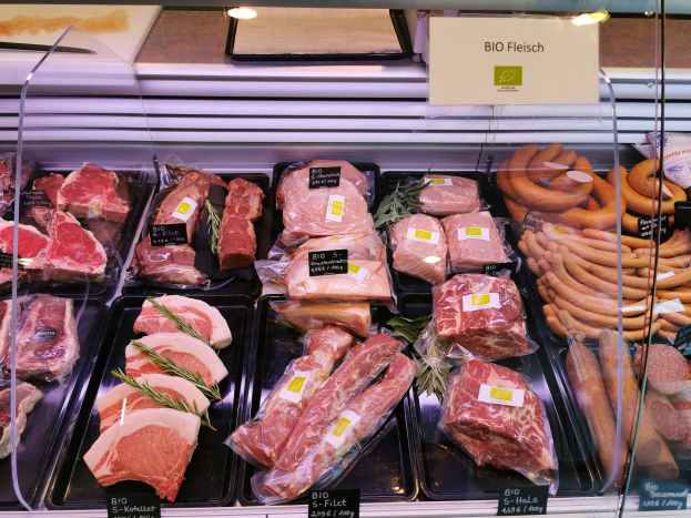 Eine Vitrine in einer Metzgerei mit verschiedenen Fleisch- und Wurstsorten in Schalen, jeweils mit Preisschildern, und einer Texttafel im Hintergrund.