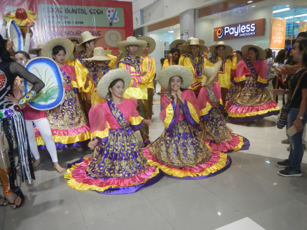 Eine Gruppe von Menschen in farbenfrohen Kleidern und Hüten, die in einem Einkaufszentrum tanzen, während andere Musikinstrumente halten und ein Banner tragen, während sie eine festliche Cinco de Mayo-Feier genießen.
