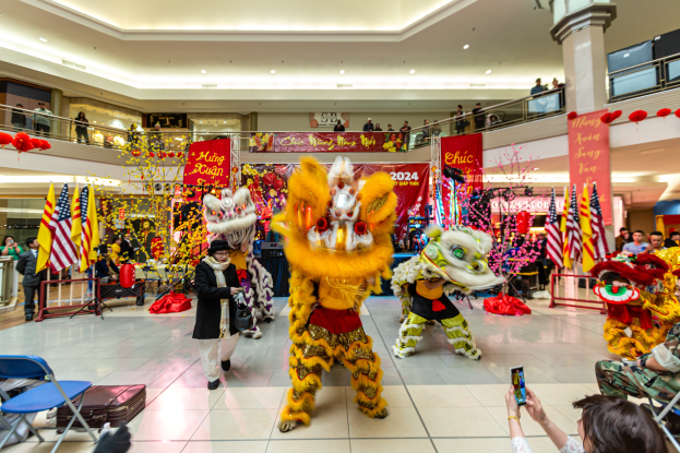 Ein lebendiges chinesisches Neujahrsfest in einem Einkaufszentrum mit tanzenden Menschen in Kostümen, anderen sitzend mit Mobiltelefonen und verschiedenen Dekorationen, Flaggen und Bannern im Hintergrund.