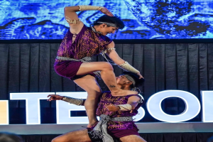 Ein Mann und eine Frau in lila Kleidern führen Akrobatik auf der Bühne durch, wobei der Mann die Frau in seinen Armen hält, während das Publikum darunter eine schwarze Gardine und einen Bildschirm sieht.