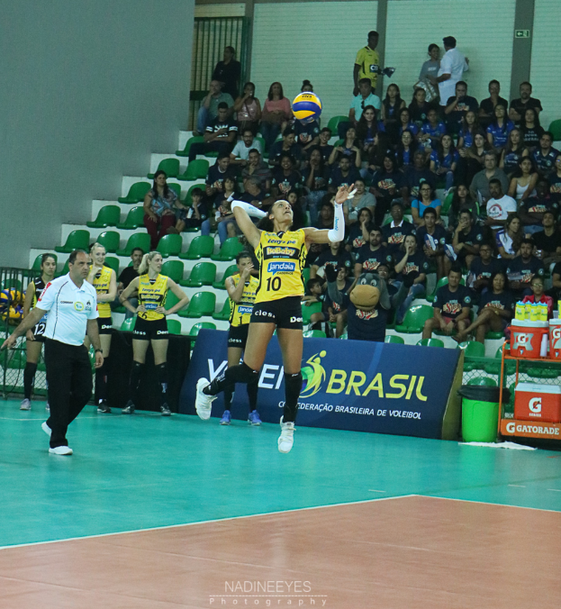 Eine Gruppe von Frauen, die Volleyball auf einem Feld spielen, mit einer Frau, die zum Schlag springt, und einem Mann auf der linken Seite, Zuschauern im Hintergrund und Text am unteren Rand des Bildes.