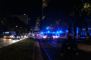Eine nächtliche Stadtstraße mit Polizei- und Busfahrzeugen in Bewegung, Fußgänger auf dem Gehweg, Gras und Bäume am Straßenrand und beleuchtete Gebäude im Hintergrund.