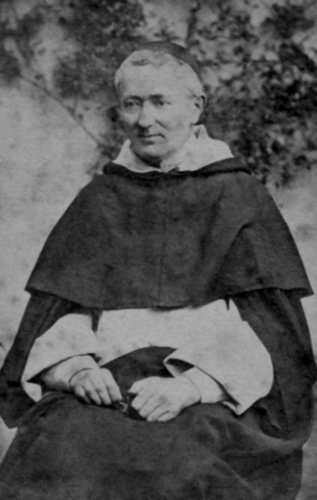 Altes Schwarz-Weiß-Foto eines Mannes in Priesterrobe, der vor Bäumen sitzt, mit schwarzer Robe, weißem Kragen und Manschetten, die Hände zum Gebet gefaltet, im warmen Licht mit friedlichem Gesichtsausdruck.