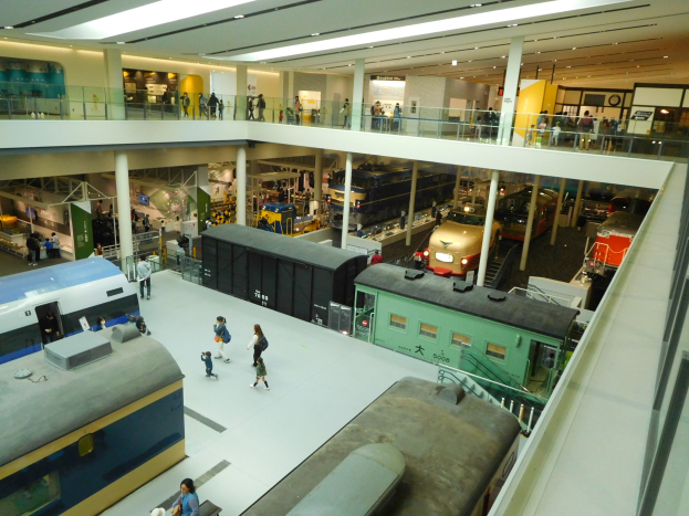 Großer Museumsraum mit verschiedenen Zugtypen, Menschen, die Ausstellungsstücke betrachten, Geländer, Säulen, Deckenleuchten und Informationsschilder.