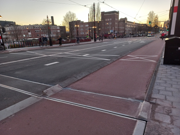 Eine Stadtstraße mit einem Fahrradweg, Fahrzeugen, Radfahrern, Laternenmasten, Bäumen, Gebäuden mit Fenstern und einem Geländer, unter einem sichtbaren Himmel.