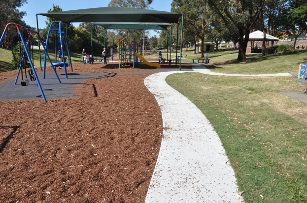 Ein Park mit einem Spielplatz, der über Schaukeln, Rutschen und andere Spielgeräte verfügt und von grünem Gras, Bäumen, Bänken, Schuppen im Hintergrund und einem klaren blauen Himmel umgeben ist.