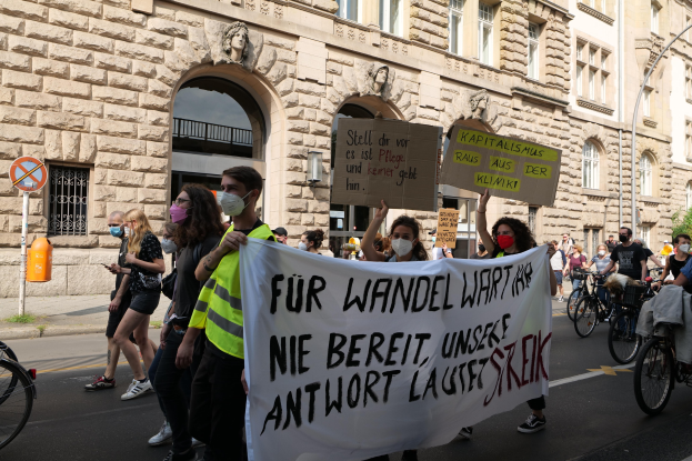 Eine Gruppe von Menschen marschiert auf einer Demonstration durch eine Stadtstraße, einige halten Schilder und andere fahren Fahrräder, im Hintergrund ein historisches Gebäude mit Rundbogenfenstern, Säulen und Skulpturen.