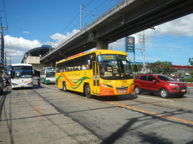 Ein gelber Bus fährt auf einer Straße neben einem roten Auto, mit Fußgängern auf dem Gehweg, Strommasten, einer Brücke, Gebäuden, Bäumen und einem bewölkten Himmel im Hintergrund.