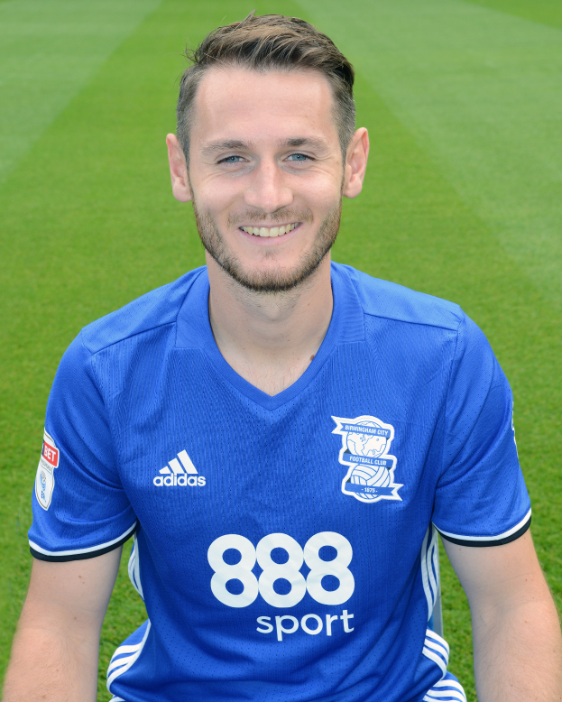 Ein Mann in einem blauen T-Shirt lächelt, während er auf einem Fußballfeld während eines Trainings sitzt.
