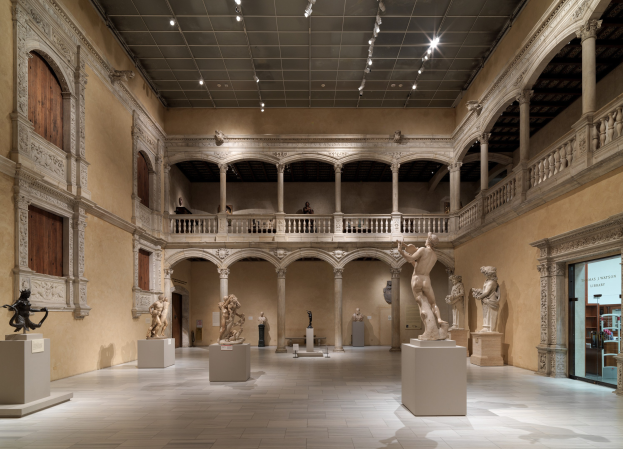 Großer Raum in einem Museum mit zahlreichen Statuen und Skulpturen auf Sockeln, architektonischen Elementen einschließlich Wänden, Säulen, Geländern und einer Decke mit Lampen, mit einigen wenigen Menschen.