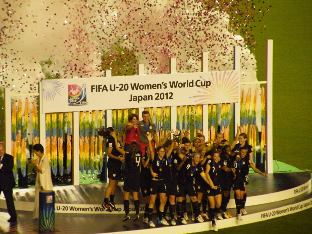 Schwarztrikotierte Frauenfußballspielerinnen feiern auf einer Bühne mit Publikum und Hintergrundanzeige.