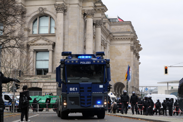 Gruppe von Polizeibeamten vor einem großen Gebäude mit Fahrzeugen auf der Straße, eine Person mit einer Kamera auf der linken Seite, Bäume und Verkehrszeichen im Hintergrund unter einem klaren blauen Himmel.