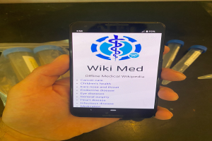 Eine Person hält ein Handy, auf dem die WikiMed-App zu sehen ist, umgeben von Reagenzgläsern und anderen Gegenständen auf dem Tisch.