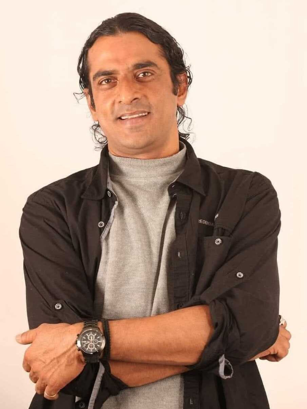 Ein Mann in einem schwarzen und aschfarbenen Kleid mit einer Uhr steht mit verschräckten Armen vor einem weißen Hintergrund.