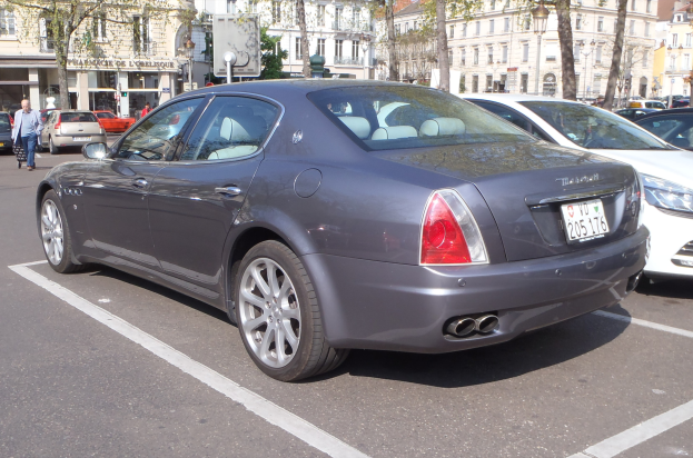 Ein Maserati Quattroporte S Q4 Gransport f├Ąhrt auf einer Stra├če mit Menschen, B├Ąumen, Geb├Ąuden mit Fenstern, Laternen und einem klaren blauen Himmel.