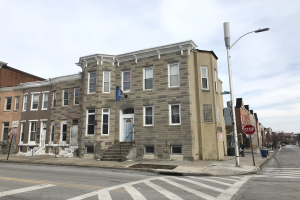 Eine Häuserreihe an einer Straßenecke in einem neuen Wohnprojekt in Philadelphia, mit Fenstern, Türen, Geländern, Laternen, Schildern und Fahrzeugen, sowie Bäumen und bewölktem Himmel im Hintergrund.