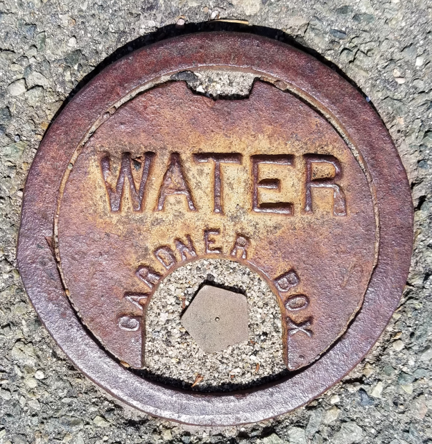 Ein verrosteter Metall-Schachtobel auf dem Boden mit dem Wort "Wasser" darauf.