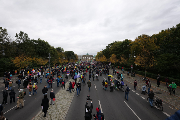 Eine große Gruppe von Menschen geht eine von Bäumen gesäumte Straße mit Laternenpfählen entlang, im Hintergrund ein Gebäude und ein klarer Himmel, einige halten Kameras während einer Protestdemo in Berlin.