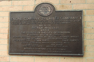 Ein rechteckiges Schild an einer Backsteinwand mit schwarzem Hintergrund und weißer fettgedruckter Schrift, die "Engine Company 2 Ladder Company" lautet.