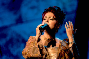 Eine Frau in einem goldenen Kleid singt auf der Bühne der Eurovision in ein Mikrofon, mit einem blauen und schwarzen Hintergrund.