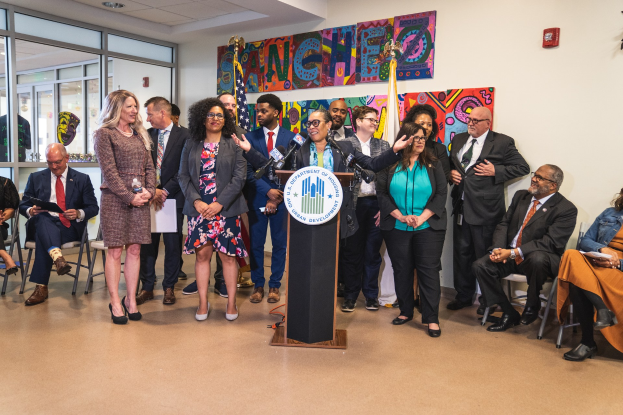 Gruppe von Menschen vor einem Rednerpult mit einem Mikrofon und einer Tafel mit der Aufschrift "Sancho City Councilman Joins City Council", mit sitzenden und stehenden Teilnehmern, Flaggen, Feuerlöscher und Deckenleuchten im Hintergrund.