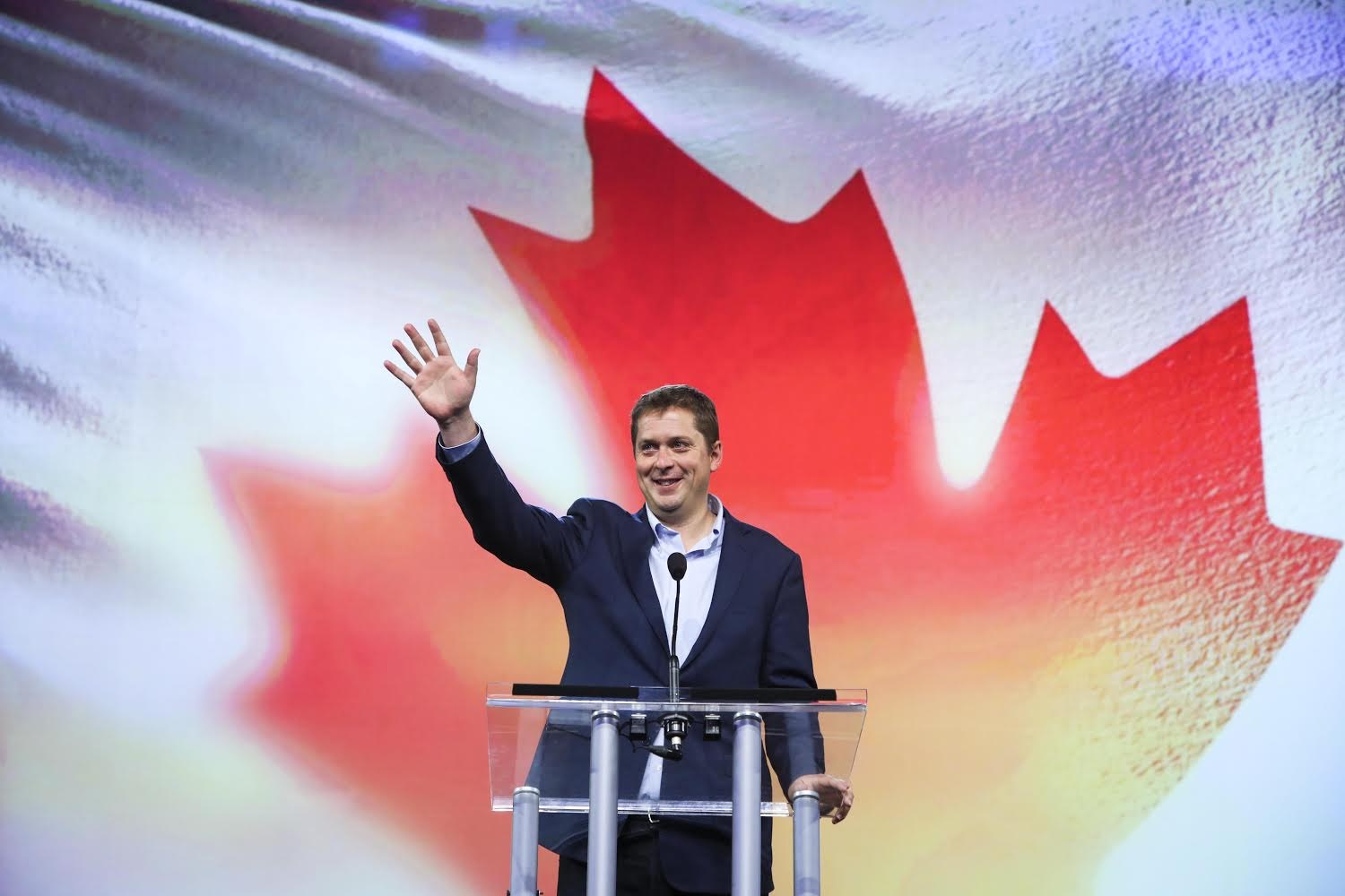 Der kanadische Premierminister Alex Salmond steht an einem Rednerpult mit Mikrofonen, lachend vor einer kanadischen Flagge, mit einem Bildschirm im Hintergrund.