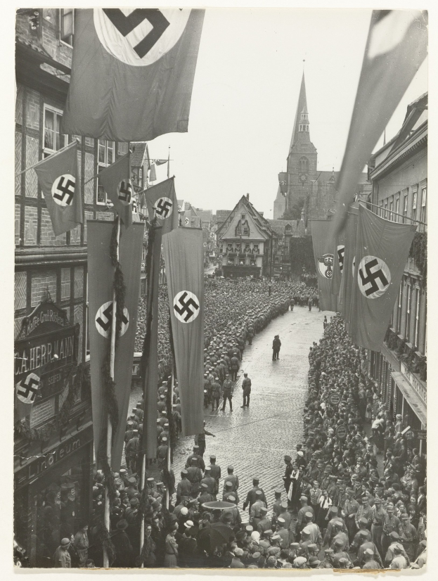 Schwarzes und wei├čes Foto einer gro├čen Menge, die eine Stra├če entlangmarschiert und Nazi-Flaggen schwenkt, mit Geb├Ąuden und einem klaren Himmel im Hintergrund.