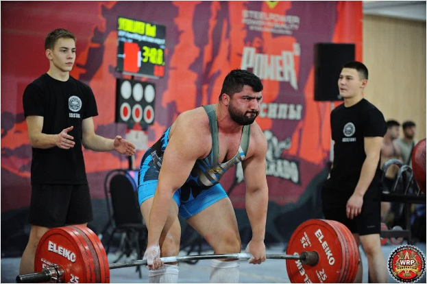 Ein Mann hebt eine Hantelstange bei einem Gewichtheben-Wettbewerb, mit Zuschauern und Equipment im Hintergrund bei den 2018 World Weightlifting Championships.