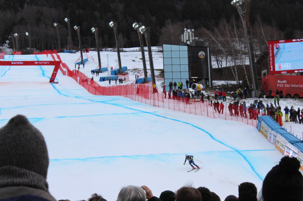 Eine Person fährt Ski auf einer schneebedeckten Abfahrt vor einer Zuschauermenge, mit einem Zaun, Laternenmasten, einem Bildschirm mit Text und Bildern und Bäumen im Hintergrund.