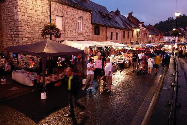 Eine Gruppe von Menschen geht auf einer Straße mit Geschäften, Schirmen, hängenden Spielzeugen, Taschen, Gebäuden mit Fenstern, Bäumen, Straßenlaternen und Barrikaden.