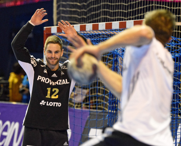 Zwei Männer spielen Handball vor einem Tor mit Netz und Banner im Hintergrund sowie einigen Zuschauern in einer Stadionumgebung.