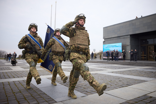 Gruppe ukrainischer Soldaten marschiert in Formation, trägt Uniformen, Helme, Handschuhe und Schuhe, hält Gewehre und Fahnen, mit Zivilisten, Pfählen, Bäumen und einem klaren blauen Himmel im Hintergrund und einer Tafel mit Text auf der rechten Seite.