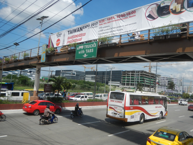 Eine belebte Stadtstraße mit Autos, Bussen, Motorrädern, einer Brücke mit Geländer und einem Banner, Laternenmasten, Strommasten mit Drähten, Bäumen, Gebäuden und einem bewölkten Himmel im Hintergrund.