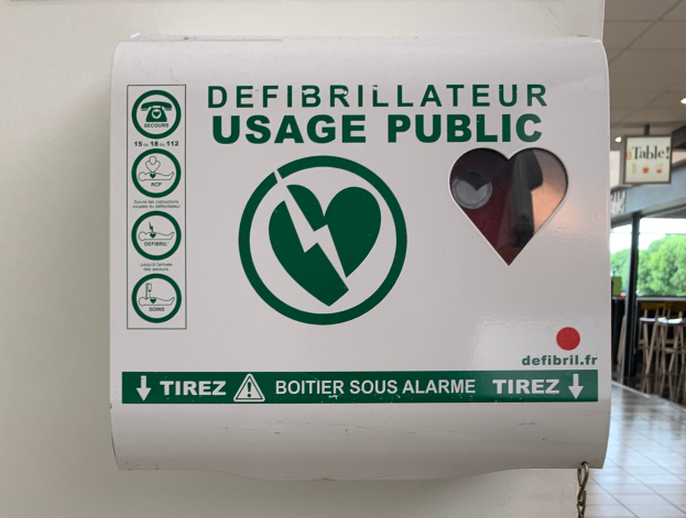Ein Defibrillator-Gebrauchs-Schild an einer Gebäudewand, umgeben von Stühlen, Tischen, Brettern, Lampen, Bäumen im Hintergrund und dem Boden unten.