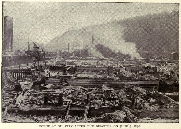 Schwarzes und weißes Foto einer Stadt nach einer Katastrophe am 5. Juni 1892, das Gebäude, Masten, Rauch, einen Hügel im Hintergrund und Text am unteren Rand zeigt.