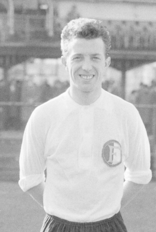 Ein Schwarz-Weiß-Foto eines grinsenden Mannes in T-Shirt und Shorts auf einem Fußballfeld mit einem Gebäude im Hintergrund, der mutmaßlich Mario Mandžukić ist.
